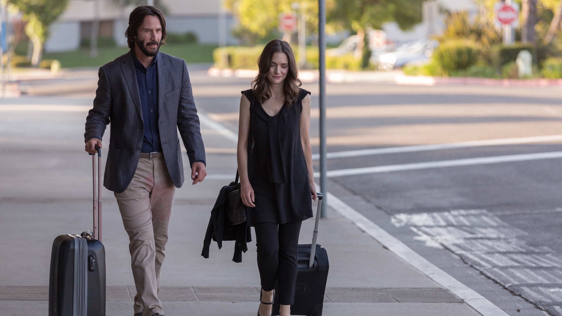 DESTINATION WEDDING: Winona Ryder und Keanu Reeves gehen nebeneinander auf einem Bürgersteig. Sie ziehen Rollkoffer hinter sich her.