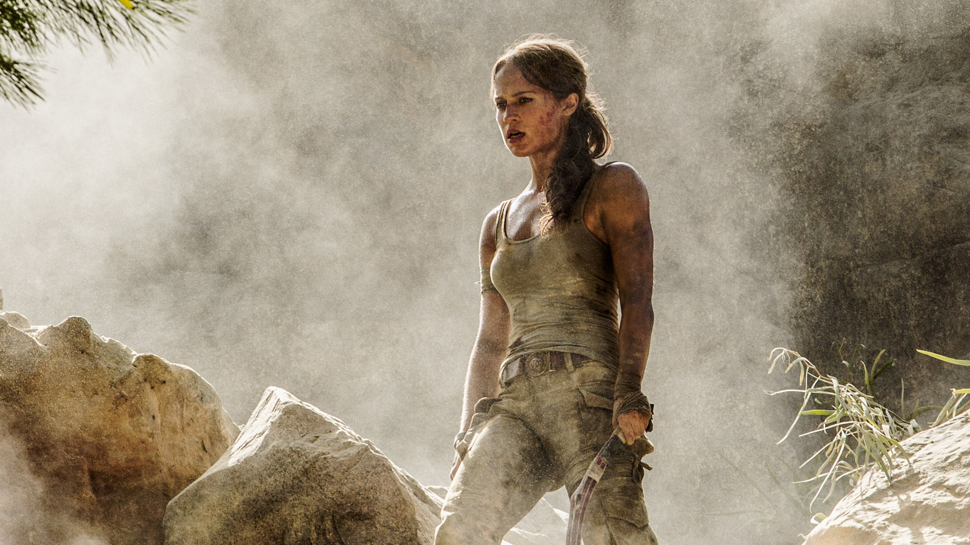 TOMB RAIDER: Lara Croft (Alicia Vikander) steht in verschmutzter Kleidung auf einem Berg. Hinter ihr steigt eine Rauchwolke auf.