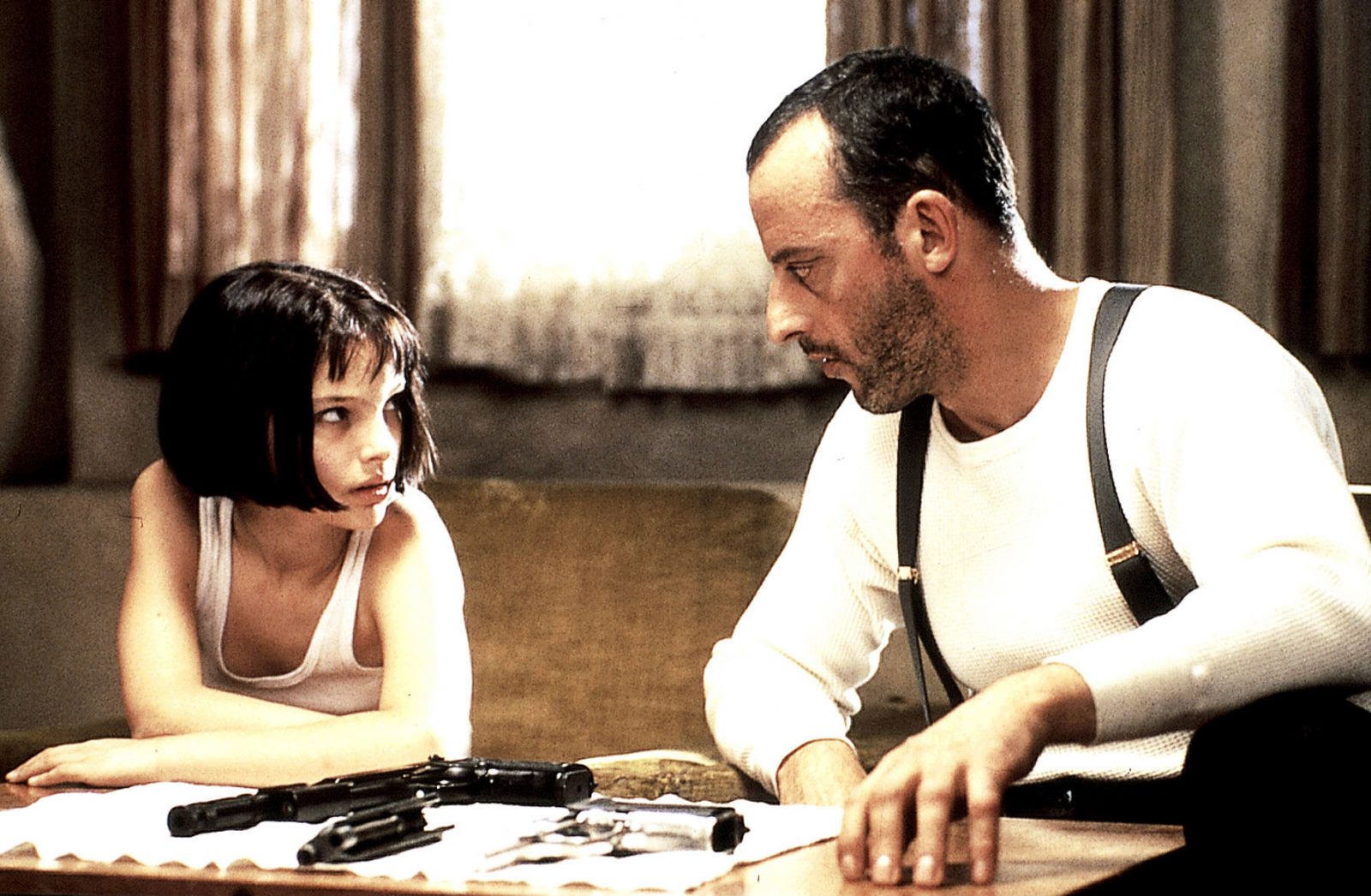 LÉON – DER PROFI: Léon (Jean Reno) sitzt neben Matilda (Natalie Portman) an einem Tisch, auf dem zwei Pistolen liegen.