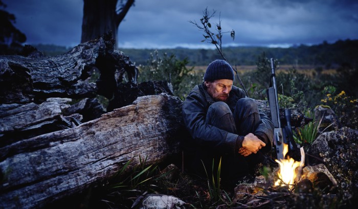 THE HUNTER: Willem Dafoe sitzt in der Dämmerung nachdenklich mit verschränkten Armen vor einem Lagerfeuer in der tasmanischen Wildnis.