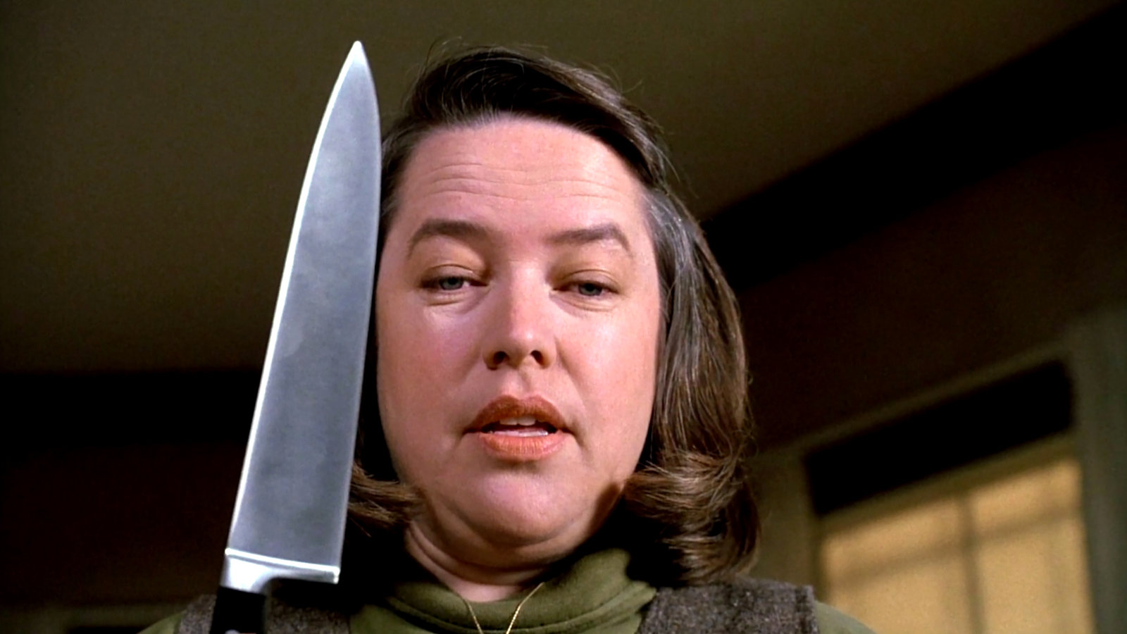 MISERY: Annie Wilkes (Kathy Bates) steht in einem verdunkelten ZImmer, hält ein großes Küchenmesser in der Hand und schaut bedrohlich herab.