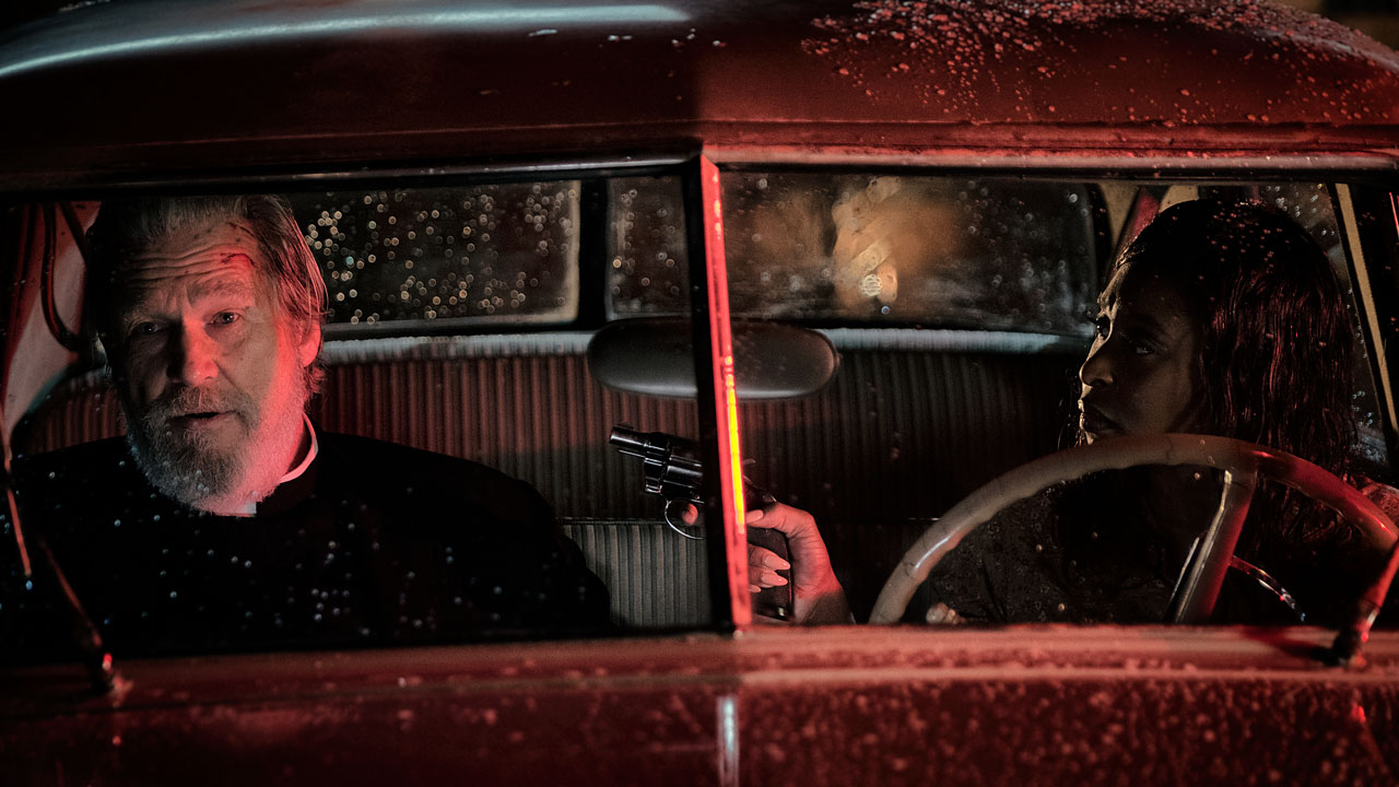 Szenenbild aus dem Kammerspiel BAD TIMES AT THE EL ROYALE: Daniel Flynn (Jeff Bridges) sitzt des Nächtens auf dem Beifahrersitz eines roten Autos. Auf dem Fahrersitz sitzt Darlene Sweet (Cynthia Erivo) und richtet eine Waffe auf ihn.