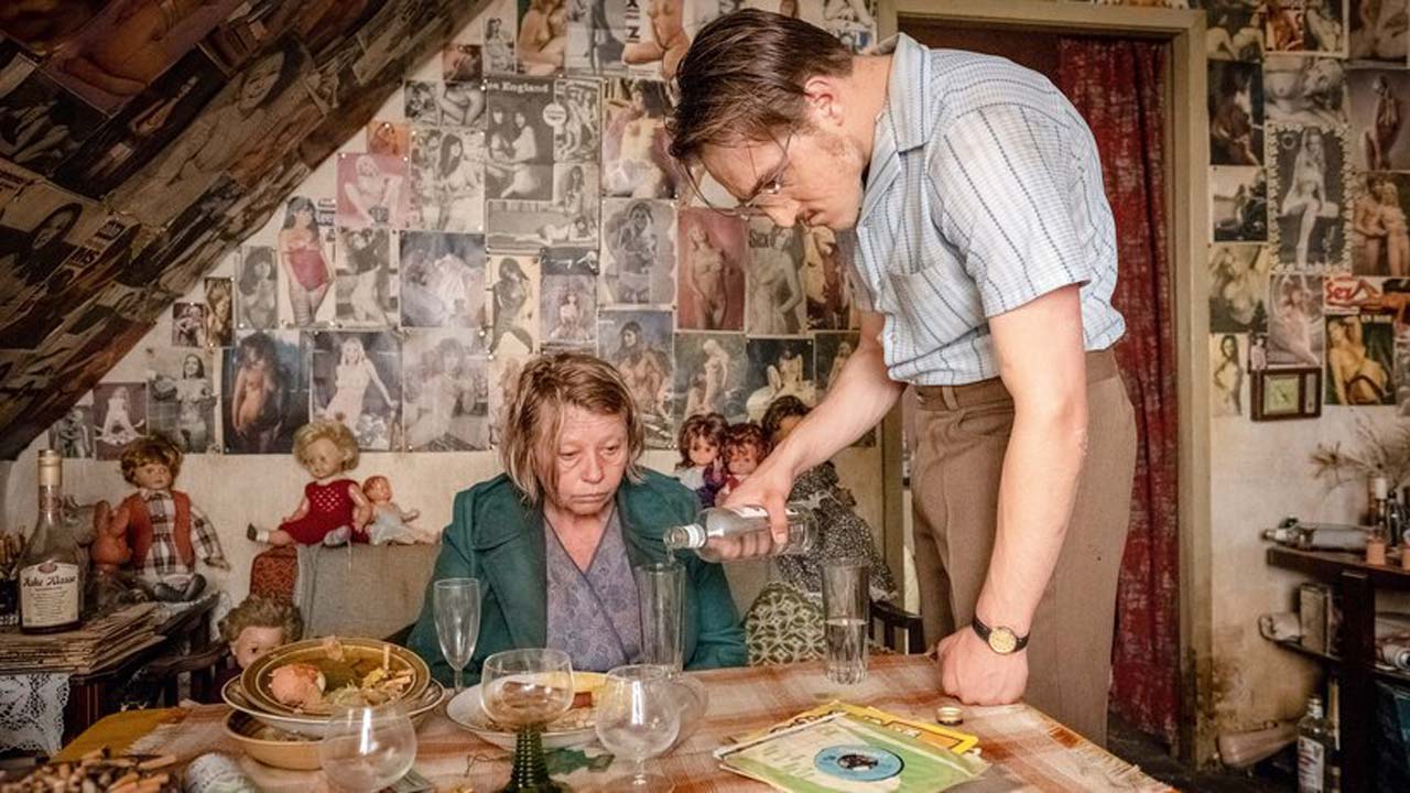 Szenenfoto aus DER GOLDENE HANDSCHUH: Fritz „Fiete“ Honka (Jonas Dassler) steht in einer Küche und schenkt der an einem Küchentisch sitzenden Gerda Voss (Margarethe Tiesel) einen Schnaps ein.