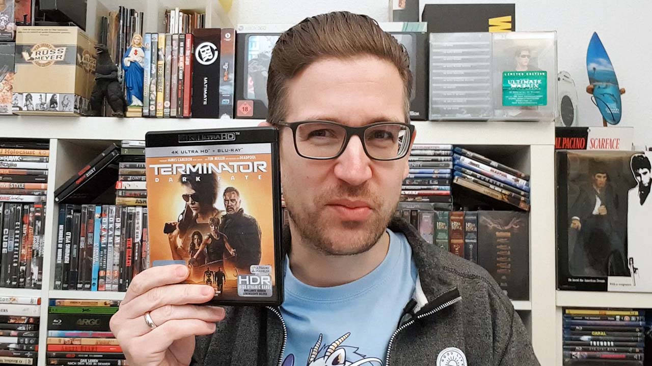 TERMINATOR – DARK FATE: Daniel Schröckert steht vor seiner Blu-ray-/DVD-Sammlung und hält lächelnd die 4K-Blu-ray-Version des Science-Fiction-Actioners mit Arnold Schwarzenegger in die Kamera.