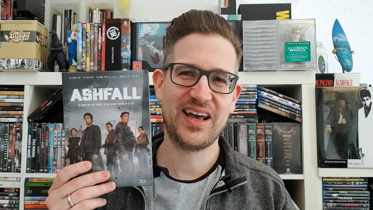 ASHFALL Review: Daniel Schröckert steht vor seinem Filmregal und hält das Mediabook des koreanischen Katastrophenfilms mit der rechten Hand hoch.
