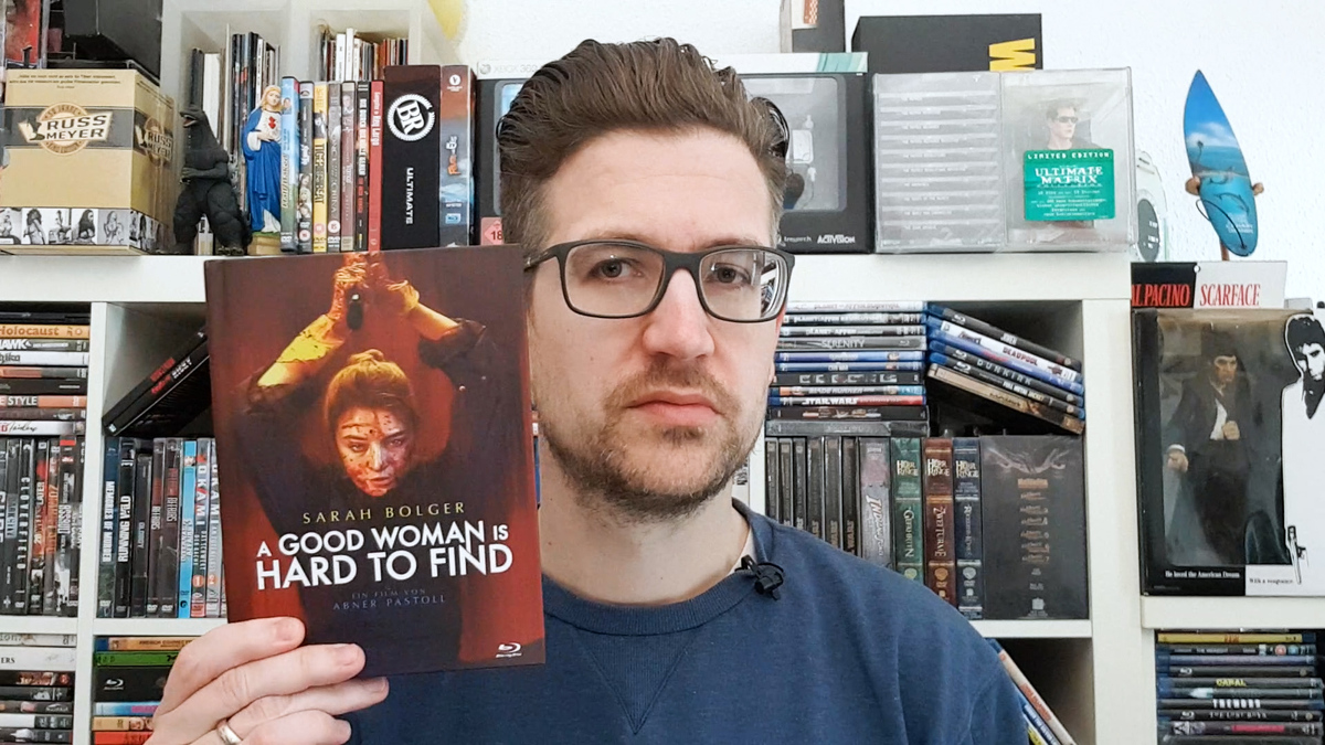 A GOOD WOMAN IS HARD TO FIND: Daniel Schröckert steht vor seinem Filmregal und hält das Mediabook des Thriller-Dramas mit der rechten Hand hoch.